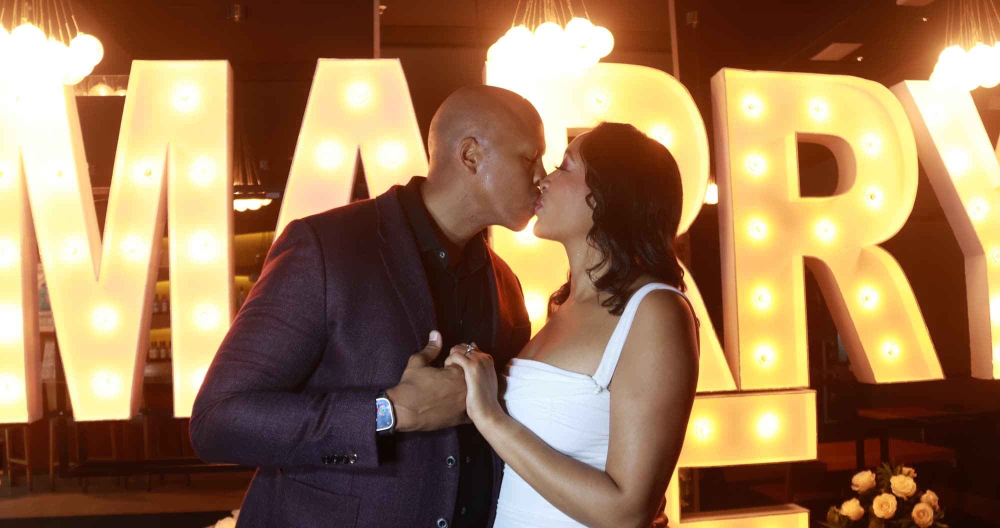 Breanna and Rob kissing in front of a lit Marry Me sign