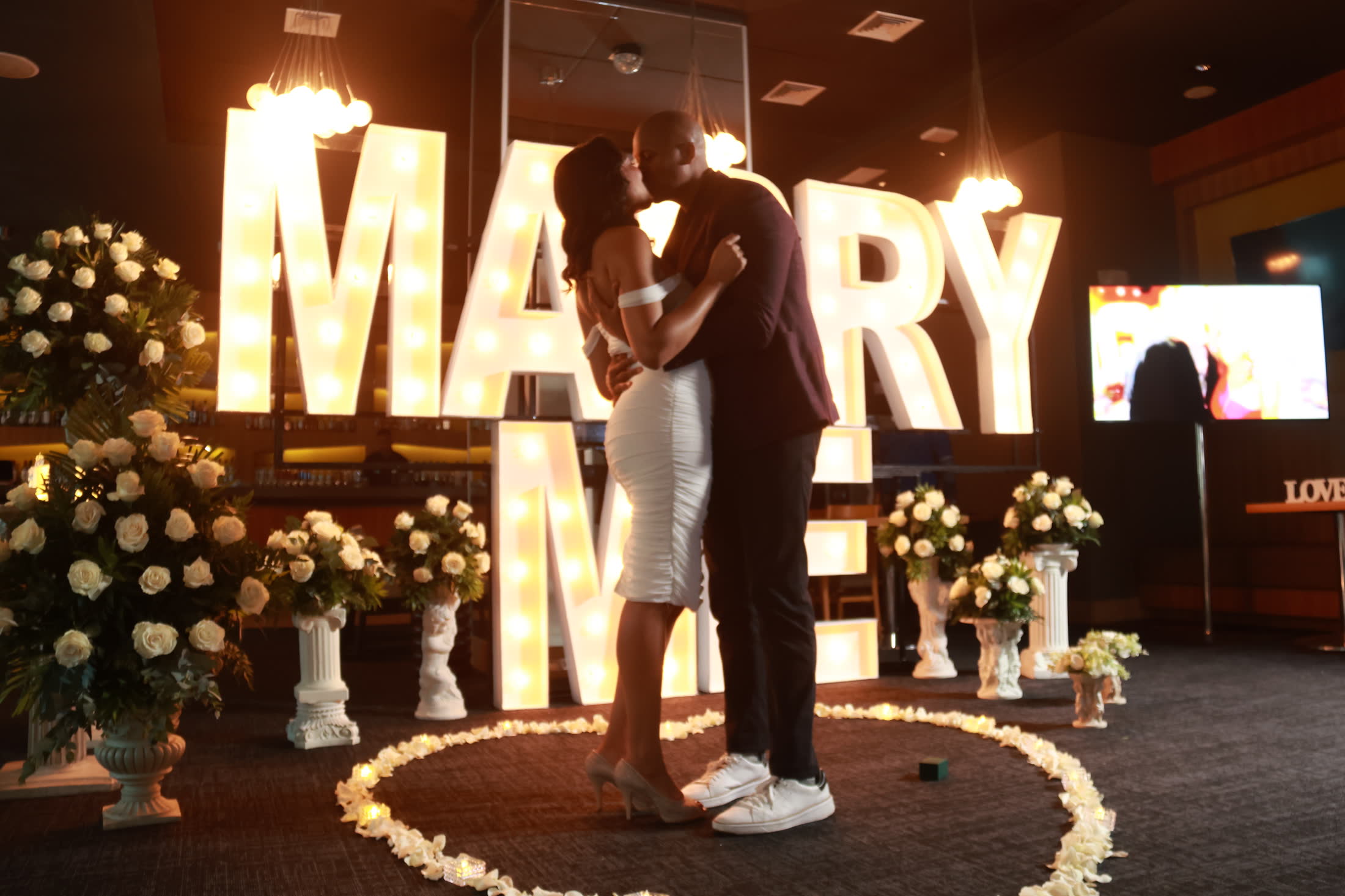 Breanna and Rob kissing under a lit Marry Me sign surrounded by white roses